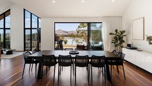 Un comedor amplio con una mesa y sillas grandes, ventanas extensas que ofrecen vistas a un lago y colinas, y un diseño interior moderno.