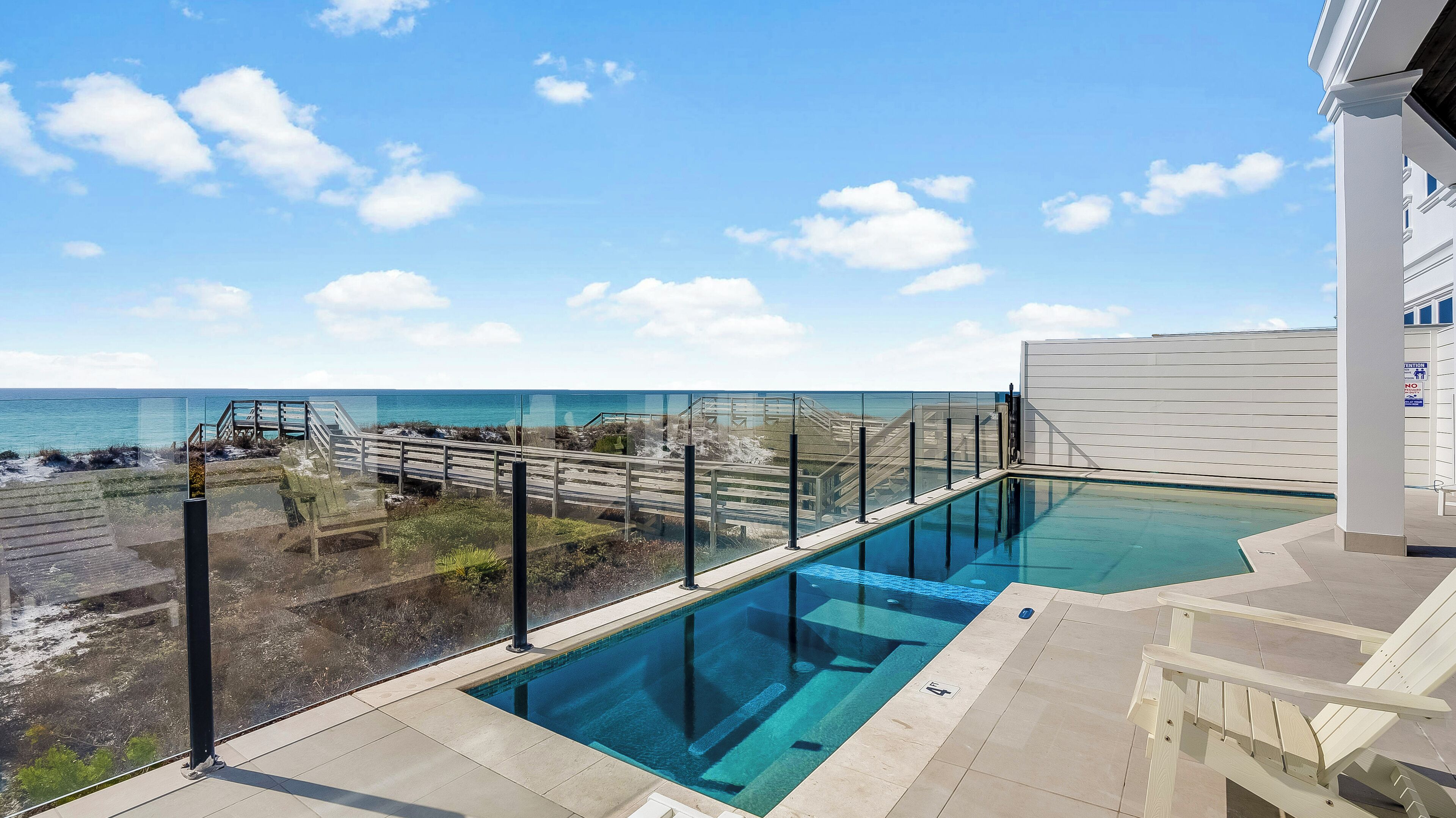 A rooftop infinity pool with a glass barrier overlooking the ocean, a wooden deck, and a clear sky.