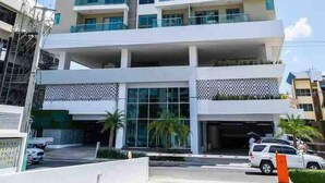 A multi-story hotel with a modern design, featuring balconies on each floor, a covered entrance, and a parking area with cars.