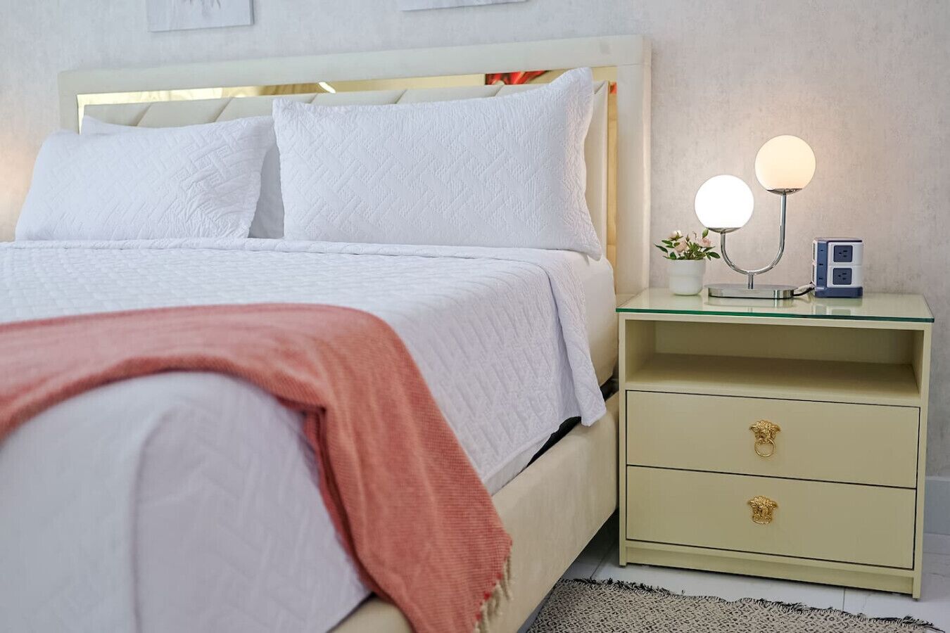 A neatly made bed with white linens and a pink throw blanket. There is a bedside table with a lamp and a small plant. The room has a simple, clean design with a light-colored wall and a small rug on the floor.