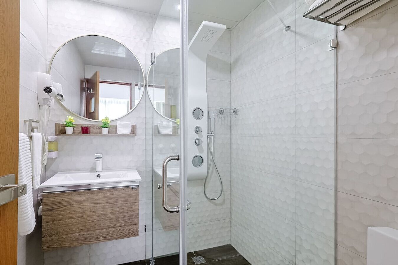 Un baño moderno con cabina de ducha de vidrio, tocador de madera, espejo redondo y una planta pequeña en el tocador.