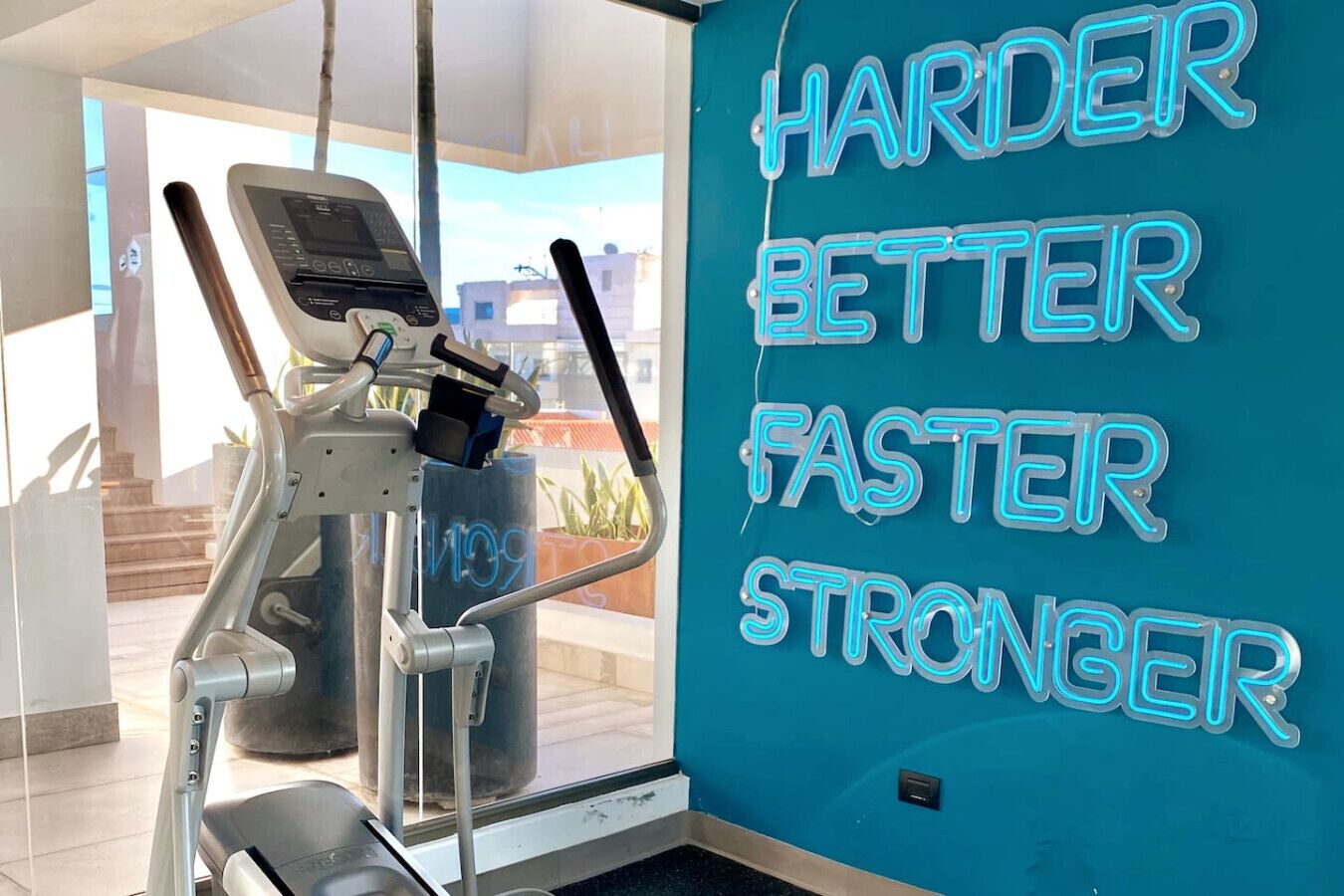 Academia com esteira e parede com letreiros de neon motivacionais dizendo 'HARDER BETTER FASTER STRONGER'.