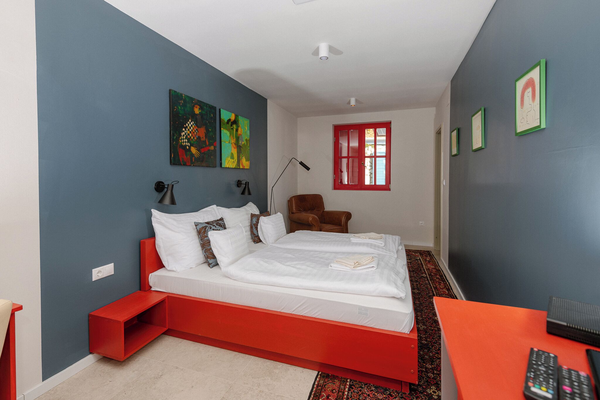 Un dormitorio moderno con un cabecero rojo, una cama individual, una mesita de noche, una silla, un escritorio y una ventana con detalles en rojo.