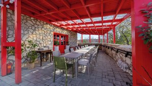 Une terrasse extérieure couverte, avec des murs en pierre, des poutres en bois rouge et une rangée de tables et de chaises.