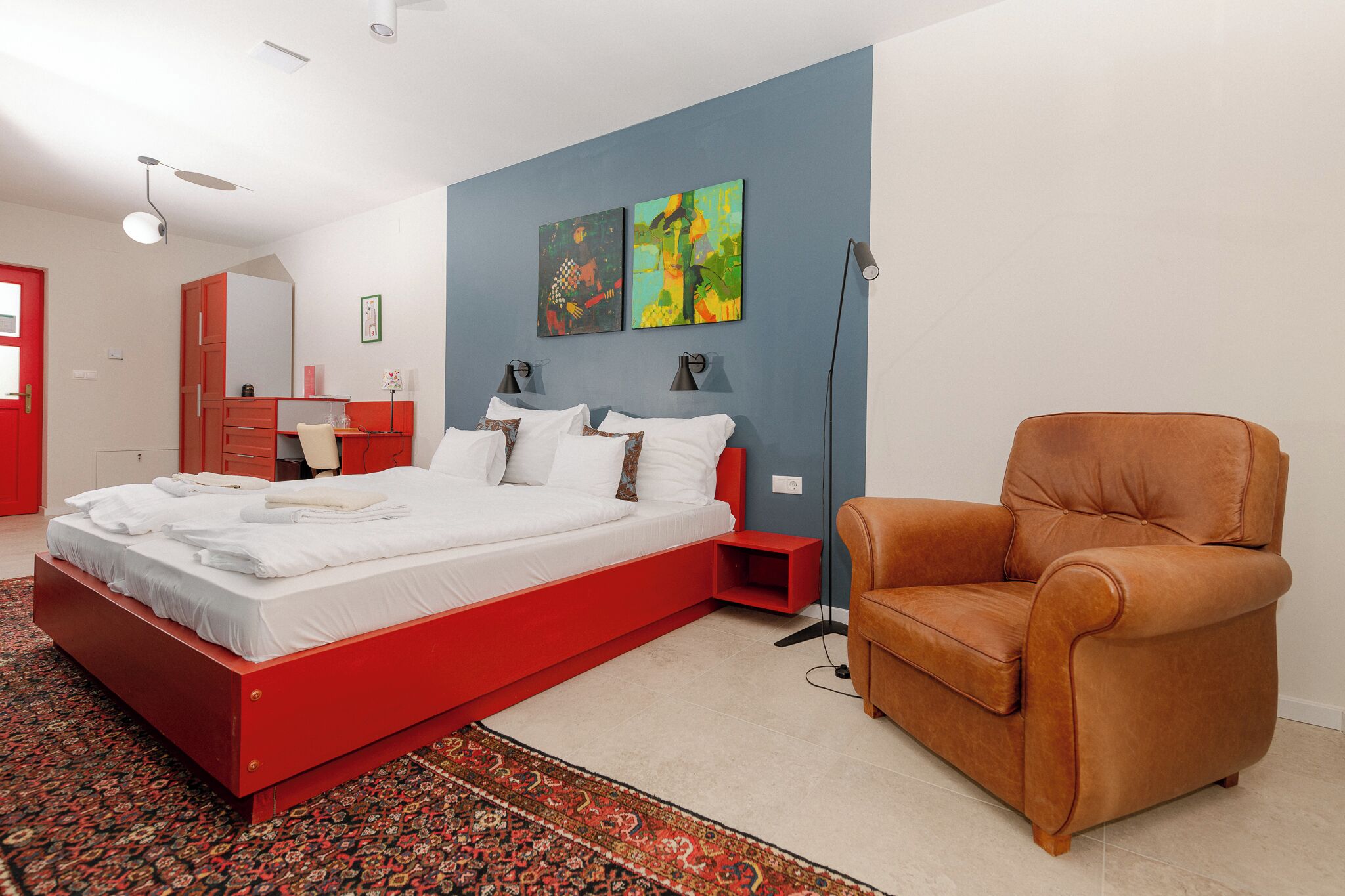 A modern bedroom with a red bed, a brown leather chair, and abstract wall art.