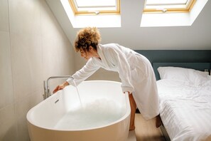 A person in a white bathrobe leaning over a freestanding bathtub in a room with a skylight and a bed with white bedding. - Bohém Borklub Panzió (Tállya)