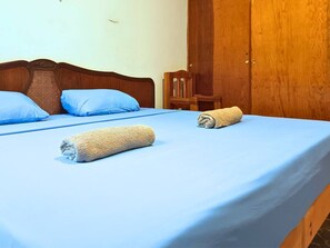 A bedroom with a wooden headboard, two blue pillows, a blue bedspread, and two rolled towels on the bed.