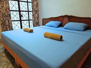 A neatly made bed with blue bedding and two rolled towels, a wooden headboard, and a window with a floral curtain.