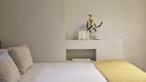 A minimalist bedroom with a bed, pillows, a bedside table with a vase of flowers, and a shelf with books. - Presidential Suites by Athens Stay (Athens)
