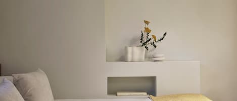 A minimalist bedroom with a bed, pillows, a bedside table with a vase of flowers, and a shelf with books.