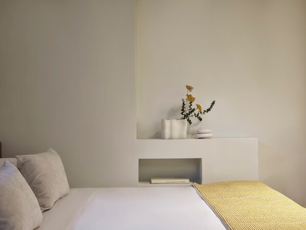 A minimalist bedroom with a bed, pillows, a bedside table with a vase of flowers, and a shelf with books.