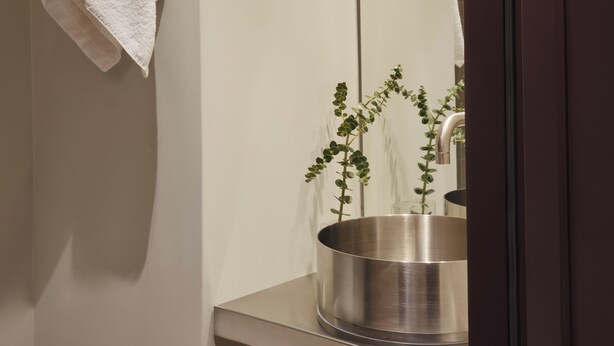 A bathroom with a towel hanging on the wall, a plant in a metallic container, and a mirror reflecting part of the room. - Presidential Suites by Athens Stay (Athens)