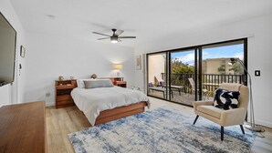 Un dormitorio con cama, mesitas de noche, un escritorio, una silla, un ventanal grande y un balcón con mobiliario exterior.