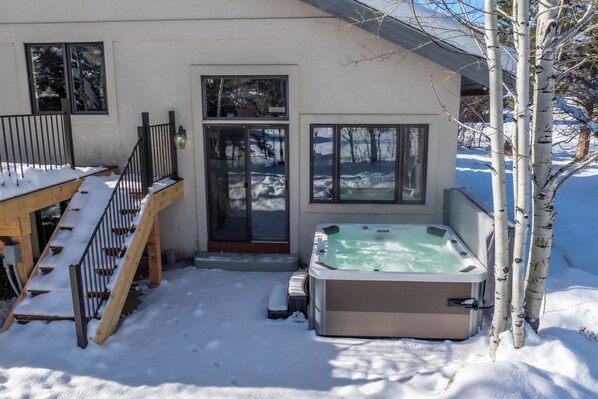 A snow-covered outdoor area with a hot tub, wooden deck, and a staircase leading to a door.