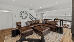 A spacious living room with a large L-shaped sectional sofa, a round wall clock, and a staircase leading to an upper level.
