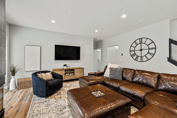 A modern living room with a sectional sofa, a flat-screen TV, and a large wall clock. - Snowbound Lodge| Private Hot Tub | Pet Friendly! (Steamboat Springs)