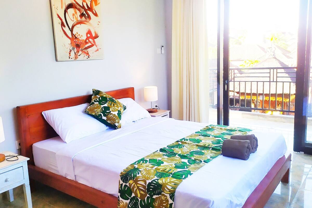 A hotel room with a bed, two bedside tables, a nightstand, a window with a view, and a painting on the wall.