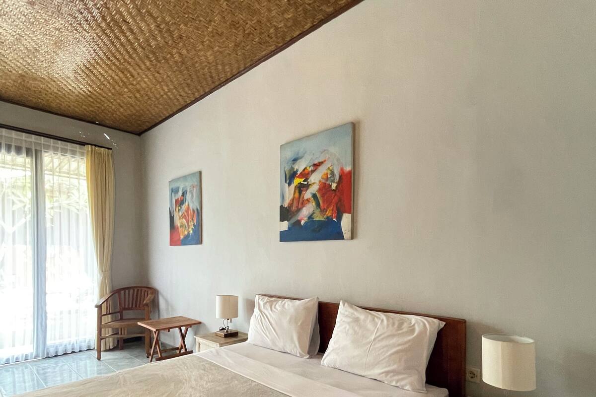 A hotel room with a large bed, two pillows, a nightstand with a lamp, a chair, and two paintings on the wall.