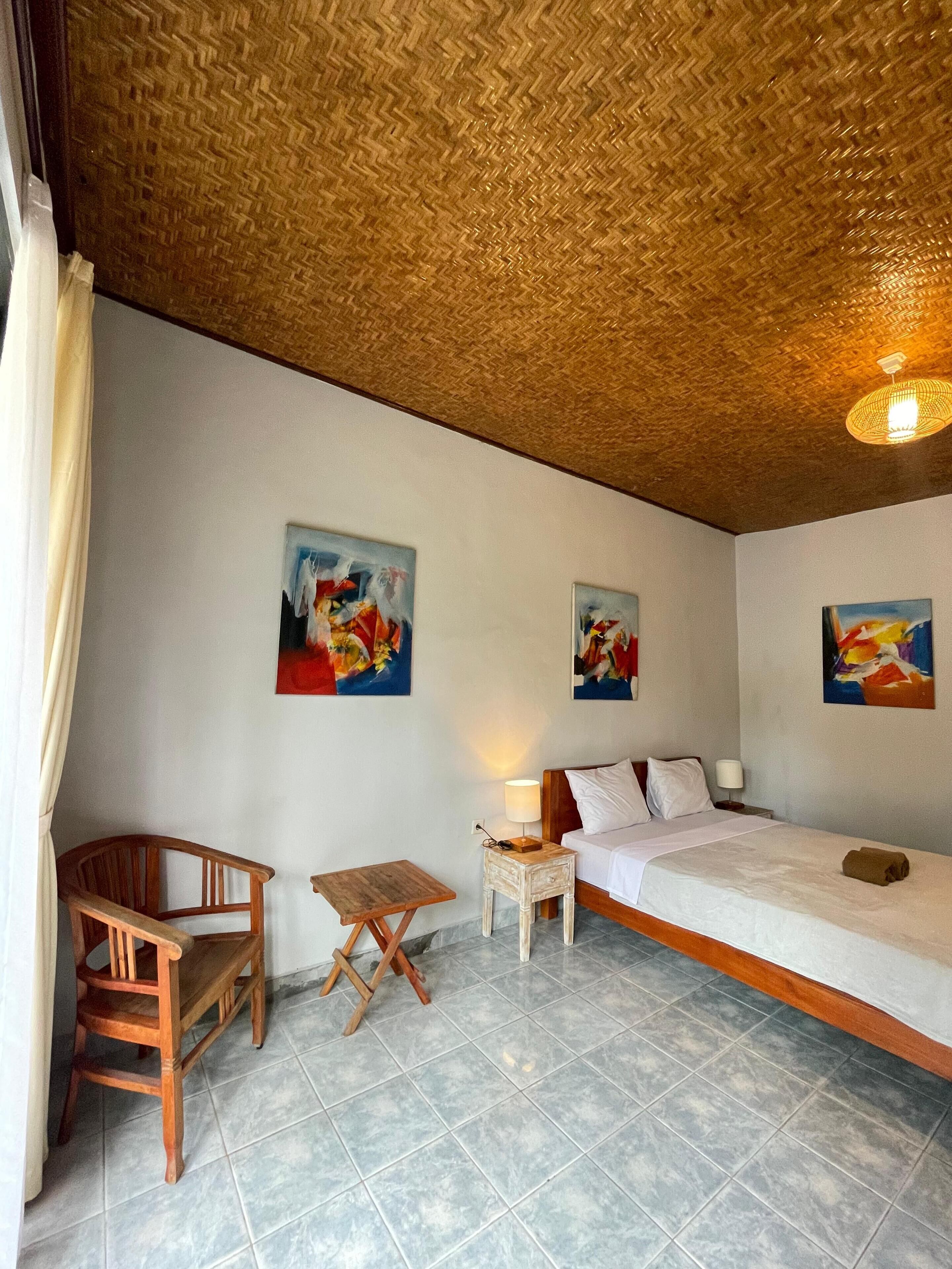 A room with a bed, a wooden chair, a small table, and two paintings on the wall.