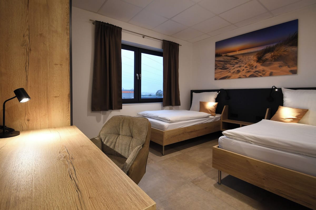 A hotel room with two beds, a wooden desk, a chair, and a large window with curtains.