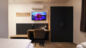 Habitación de hotel con cama, escritorio con silla, televisión montada en la pared y armario.