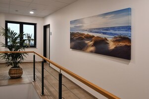 Um corredor com uma pintura grande de uma cena de praia, um vaso com planta e um corrimão de madeira.