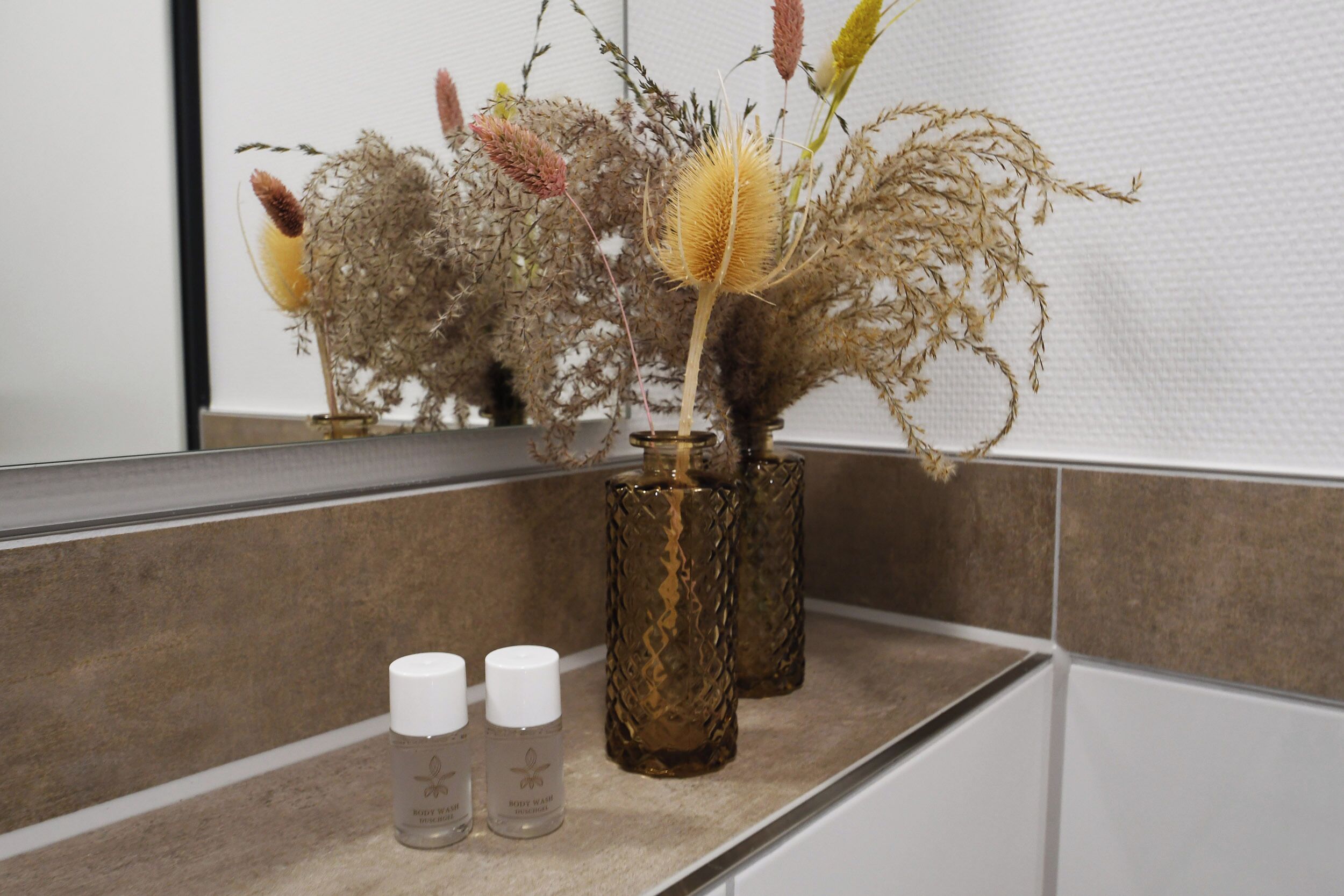 Un meuble-lavabo de salle de bain avec un miroir, un vase contenant des fleurs séchées et trois flacons de gel douche.