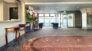 Un vestíbulo de hotel con una mesa de recepción, un gran arreglo floral, una zona de estar con sofás y vistas al exterior a través de amplias ventanas.