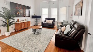 Een moderne woonkamer met een houten salontafel, een zwarte leren bank, een grijze fauteuil en een grote flatscreen-tv aan de muur bevestigd.
