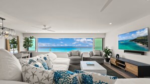 A modern living room with a large window overlooking a beach, a ceiling fan, and a flat-screen TV on the wall.