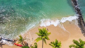 An aerial view of a beach with clear turquoise water, a sandy shore, and palm trees.