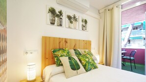 A bedroom with a wooden headboard, a bedside table with a lamp, and a bed with tropical leaf-patterned pillows. There are wall-mounted potted plants above the bed.