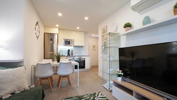 A modern living room with a flat-screen TV, a dining area with a white table and chairs, and a kitchen in the background.