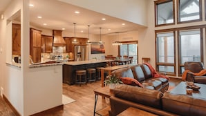 A spacious kitchen with wooden cabinets and a central island, a dining area with a brown leather couch, and large windows offering views of the outdoors. - Spacious 2-Level Custom Craftsman Style Lodge w/ Hot Tub, Fire Pit, Clubhouse (Lead)