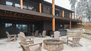 A wooden deck with Adirondack chairs and a stone fire pit, overlooking a modern building with large windows and a balcony. - Spacious 2-Level Custom Craftsman Style Lodge w/ Hot Tub, Fire Pit, Clubhouse (Lead)