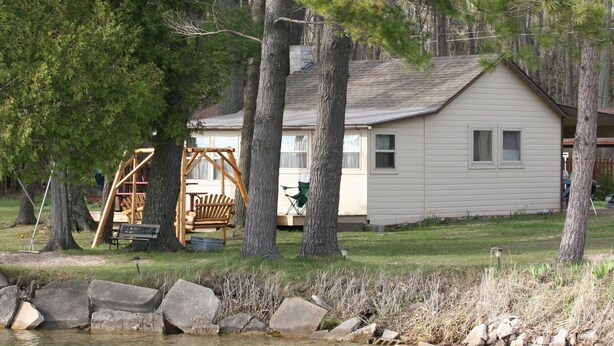 A cabin with a simple, single-story structure, surrounded by trees and a small body of water. - Affordable Family Fun on the Platte Lakes Van Hammen Cottage #3 (Honor)