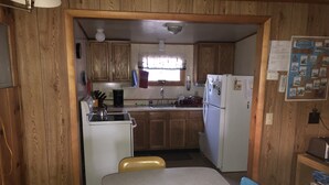 A compact kitchen with wooden cabinets, a refrigerator, a stove, and a sink. There is a dining table with chairs and a bulletin board with a map. - Affordable Family Fun on the Platte Lakes Van Hammen Cottage #3 (Honor)