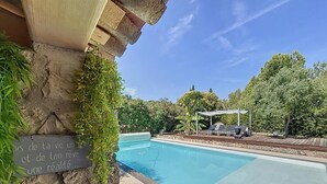 A clear blue swimming pool with a stone wall and greenery on one side, and a covered seating area with lounge chairs on the other. - Family Villa with swimming pool tennis spa (Auribeau sur siagne)