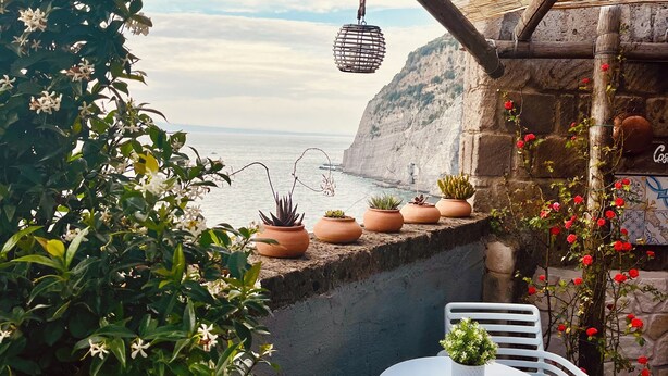 Eine Terrasse mit Meerblick, Kübelpflanzen, einem hängenden Laternen und einem Tisch mit Stühlen.