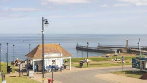 A coastal scene with a pier extending into the sea, a small building with a tiled roof, and a street with pedestrians.