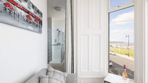 A modern bathroom with a glass shower enclosure, a white door, and a view of a coastal road through a large window.