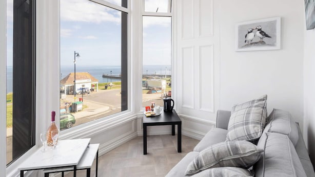A room with a large bay window, a small table with a kettle, and a grey sofa.