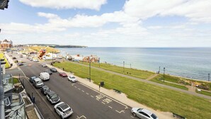 Uma estrada costeira com carros estacionados, uma área gramada e vista para o mar.