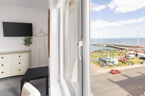 A room with a view of a coastal area, a TV mounted on the wall, a desk, and a chair.