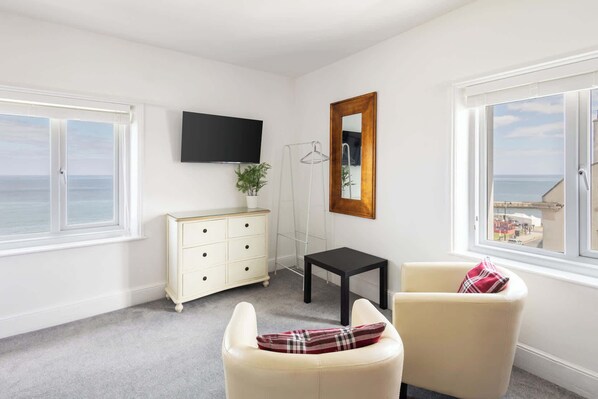 A room with a white dresser, a black table, and two chairs with a plaid pillow. A TV is mounted on the wall, and there is a window with a view of the sea.
