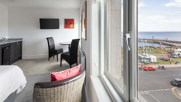 A modern hotel room with a large window, a dining area with two chairs, a flat-screen TV, and artwork on the wall.