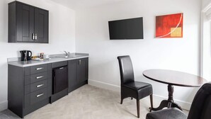 A modern kitchen with dark cabinetry, a built-in dishwasher, and a countertop with a kettle and coffee maker. There is a dining area with a round table and two chairs, and a flat-screen TV mounted on the wall.