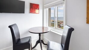 A corner room with a round table and two leather chairs, a flat-screen TV mounted on the wall, and a window with a view of the sea and buildings.