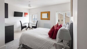 A modern bedroom with a large bed, a desk, and a chair. There is a window with blinds, a mirror, and a lamp on the bedside table.