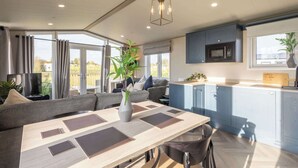 A modern living room with a sofa, a dining table with placemats, a potted plant, and a kitchen area with blue cabinets and a microwave. - Host & Stay | Riverwood, Ashdown Retreat (Pickering)